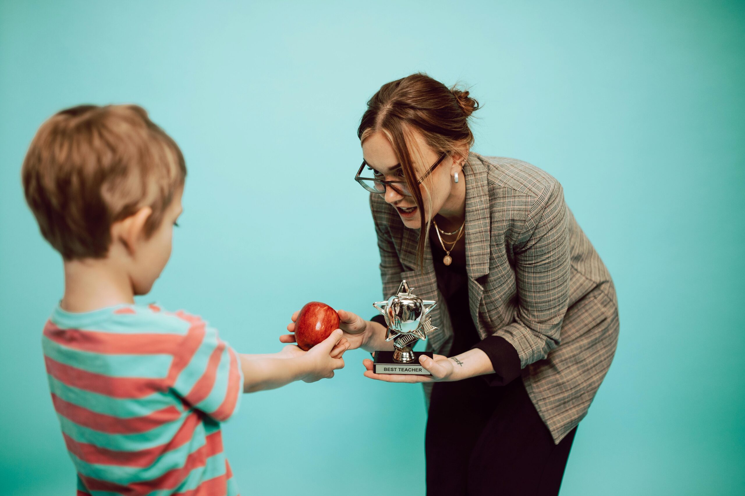 A child offers an apple to a teacher holding a "Best Teacher" trophy in a classroom scene.